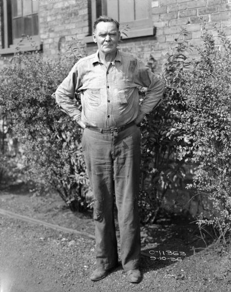 Full-length portrait of a man with his hands on his hips standing outdoors in front of bushes along the side of the brick wall of a factory building with windows. He is wearing work clothes.