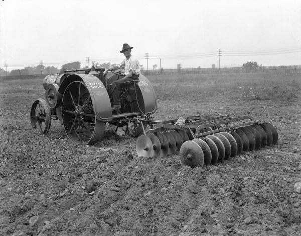 Three-quarter rear view from left of a man driving a Titan 10-20 tractor  pulling a 6 ft. disc harrow in a field.