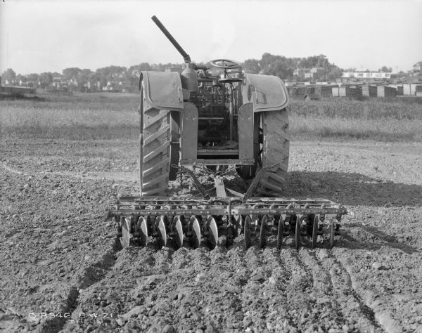 Rear view of a tractor hitched to a disc harrow in a field. In the background are stacks of lumber and buildings.