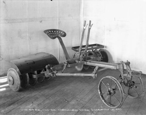 Three-quarter view from front right of an orchard disk harrow (exp) set up on a wood floor in front of a backdrop.