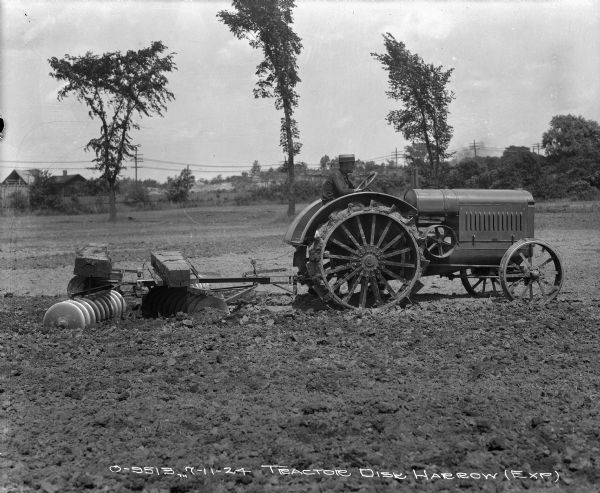 Right side profile view of a man driving a tractor pulling an experimental weighted disk harrow. Buildings are in the background.
