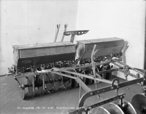 Three-quarter view from right rear of a seeding attachment on a disk harrow set up on a wood floor. There is a light-colored backdrop behind the machinery.
