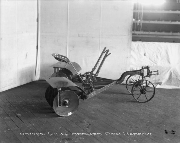 Right side view of an orchard disk harrow set up on a wooden floor in the corner of a room with a backdrop behind it.