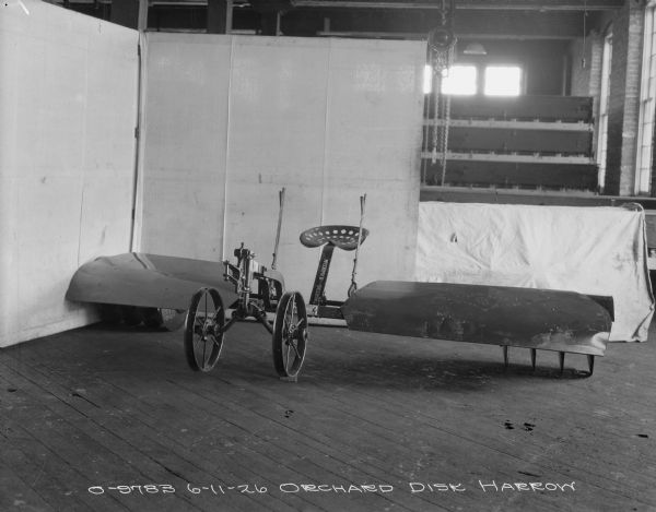 Three-quarter view from front left of an orchard disk harrow set up on a wooden floor in the corner of a room with a backdrop hanging partially behind it.