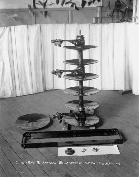 Bumper disk harrow, with parts and disks set up on the wood floor. In the background is a half-curtain backdrop. There are parts hanging on the wall in the background above the curtain.