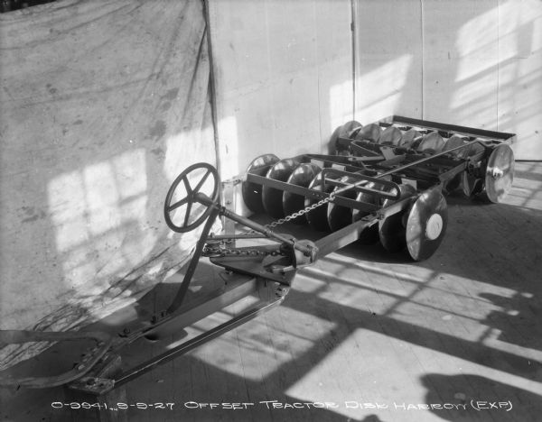 Slightly elevated view of an experimental offset tractor disk harrow set up on a wood floor near a wall and a backdrop.
