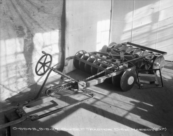 Slightly elevated view of an experimental offset tractor disk harrow set up on a wood floor near a wall and a backdrop.
