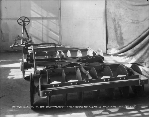 Rear view of an offset tractor disk harrow set up on a wood floor in a room with cloth backdrops in the background.
