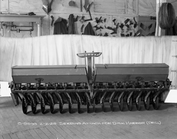 Rear view of a seeding attachment for a disk harrow (drill) set up on a wood floor. There is a half-curtain backdrop in the background. Hanging on the wall behind the backdrop are coats, jackets, and parts.