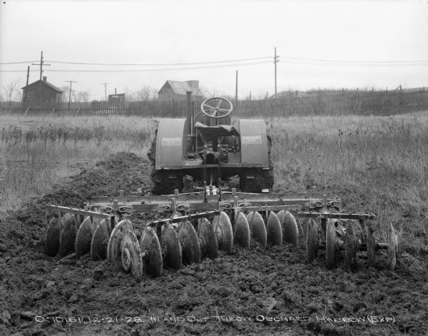 Rear view of a McCormick-Deering 10-20 Hitched to Orchard Disk Harrow sitting in a field. In the background is a fence, and beyond the fence are wood buildings and power lines.