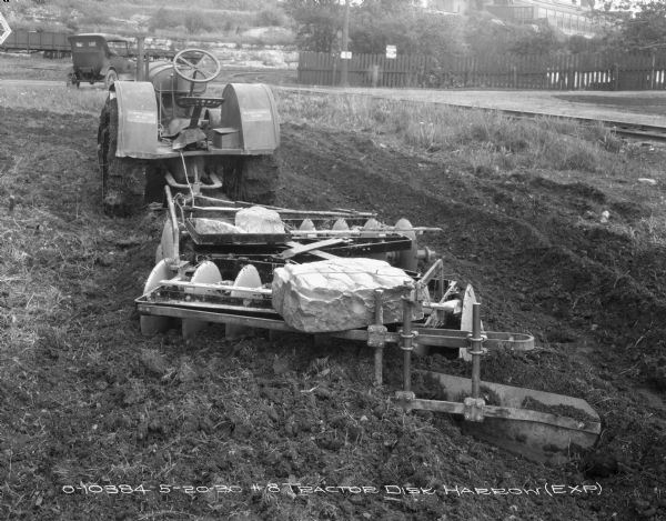 Rear view of a disk harrow in a field hitched to a McCormick-Deering 10-20 tractor. There are rocks on top of the disk harrow for extra weight. There is an automobile parked further down the field, and in the background on the left is a railroad car, and on the right is a fence. A factory building is in the far distance on a hill on the right.