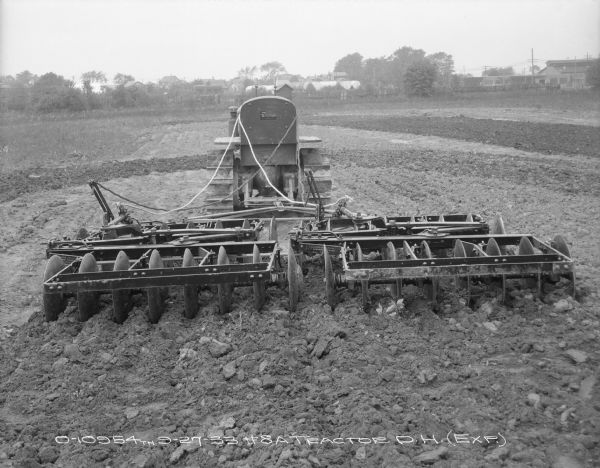 Rear view of an experimental #8A disk harrow hitched to a continuous track, TracTracTor, tractor in a field. Buildings are in the background beyond a fence.