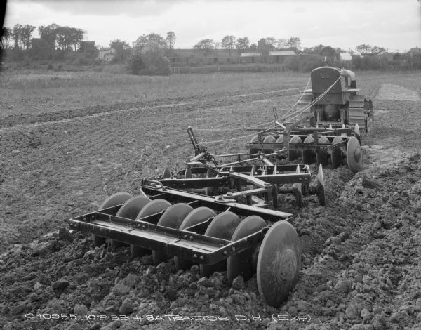 Rear view of an experimental #8A tractor disk harrow hitched to a continuous track, TracTracTor, tractor in a field. Buildings are in the background beyond a fence.