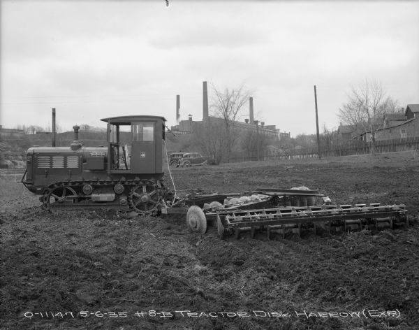 Left side view of a TracTracTor pulling an experimental #8B disk harrow in a field. There are automobiles parked in the background near a fence. On a hill in the distance is a factory building with smokestacks.
