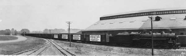 View down railroad tracks looking towards a long line of railroad cars on another set of tracks. The large banners on the side of some of the cars reads: "Train Load of International Trucks for Dealers Philadelphia Territory." A large industrial building is behind the cars. An engineer can be seen in the locomotive on the right, and the coal car behind has a sign painted on the side that reads: "New York Central Lines."