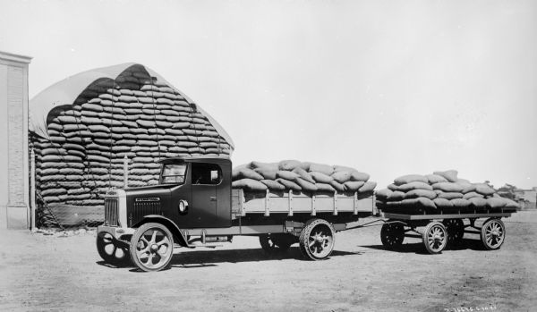 A delivery truck with a truck bed and a trailer loaded with sacks of grain. In the background behind a fence is a tall stack of more sacks covered with a tarp.