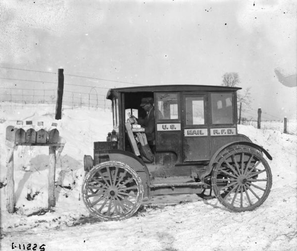 View towards a man sorting mail in a truck with a sign on the side that reads: "U.S. Mail R.F.D." There are four mailboxes on wooden posts on the left, and a fence is on a slope in the background. Snow is on the ground.