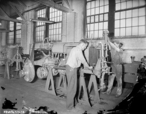 Two men are working on a assembly line, as part of steps in assembling a Farmall tractor.
