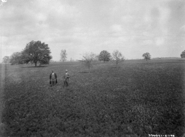 Slightly elevated view of two men wearing suits and hats standing in a field. Trees are in the background.
