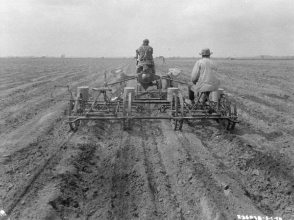 Rear view of two men working in a field. One man is sitting on the planter, and another man is driving the Farmall tractor.