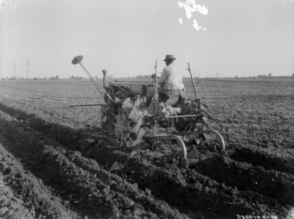 Three-quarter view from left rear of a man using a Farmall tractor to pull a cultivator in a field.