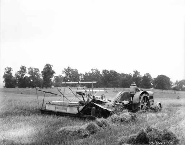 Three-quarter view from right rear of a man driving a tractor pulling a McCormick-Deering binder in a field.