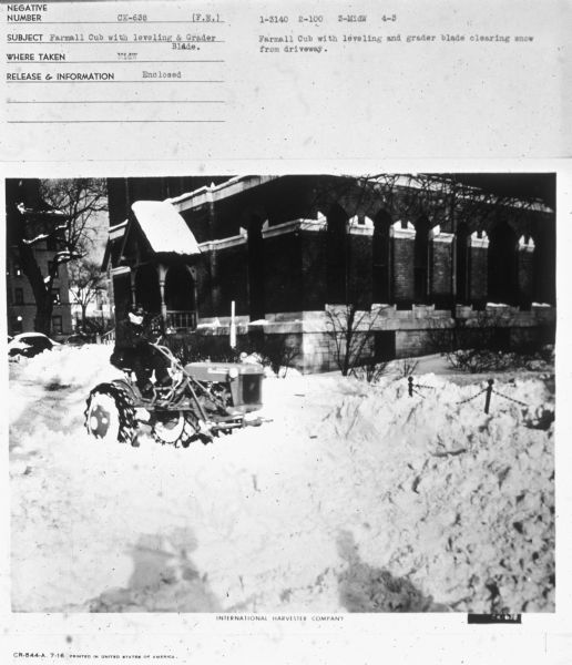 Three-quarter view from front right of a man driving a Farmall Cub to clear snow near a large brick building. Subject: "Farmall Cub with leveling & Grader Blade." Where Taken: "MidW." Information with photograph reads: "Farmall Cub with leveling and grader blade clearing snow from driveway."
