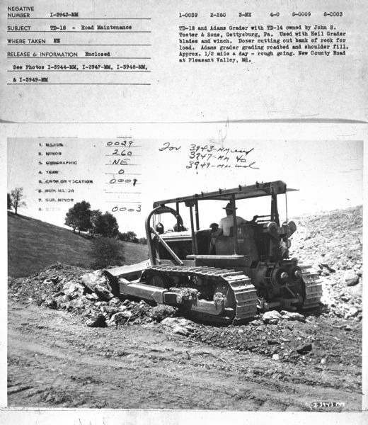 Three-quarter view from left rear of a man driving a TD-18 crawler tractor.
Subject: "TD-18 — Road Maintenance." Where Taken: "NE." Information with photograph reads: "TD-18 and Adams Grader with TD-14 owned by John S. Teeter & Sons, Gettysburg, Pa. Used with Heil Grader blades and winch. Dozer cutting out bank of rock for load. Adams grader grading roadbed and shoulder fill. Approx. 1/2 mile a day — rough going. New County Road at Pleasant Valley, Md."