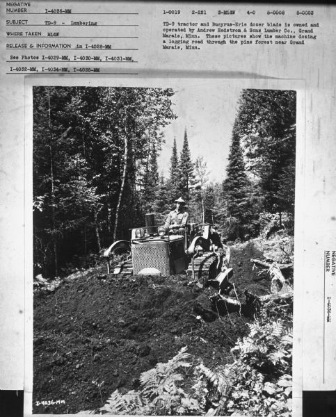 Front view of a man driving a TD-9 crawler tractor.
Subject: "TD-9 — Lumbering." Where Taken: "MidW." Information with photograph reads: "TD-9 tractor and Bucyrus-Erie dozer blade is owned and operated by Andrew Hedstrom & Sons Lumber Co., Grand Marais, Minn. These pictures show the machine dozing a logging road through the pine forest near Grand Marais, Minn."
