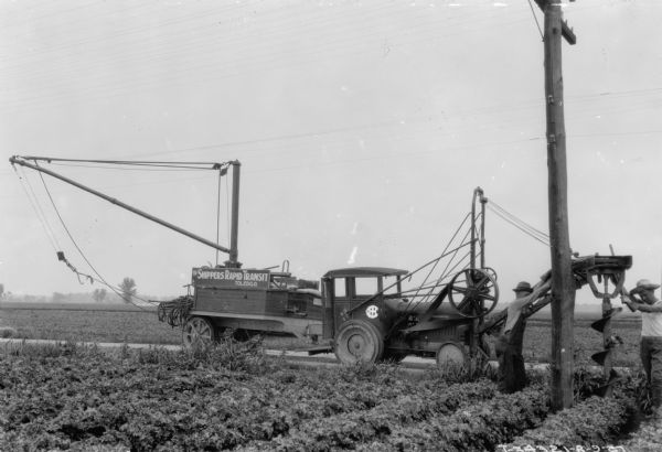 View from field towards men installing telephone poles with a large drill attached to the front of a truck. The sign on the truck reads: "The Shippers Rapid Transit."