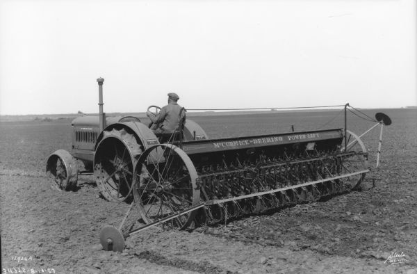 Three-quarter view from left rear of a man driving a McCormick-Deering 15-30 tractor that is pulling a power lift seeder in a field in Saskatoon, Canada.