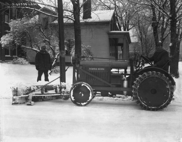 Left side view of a man driving a tractor with a snow plow on the front. He is clearing a road, and a man is standing and watching from the sidewalk on the left. Buildings and houses are in the background.
