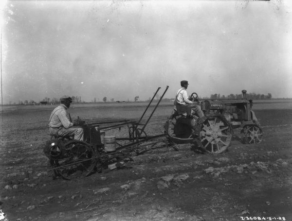 Right side view of a man driving a Farmall tractor pulling a man on a planter in a field.