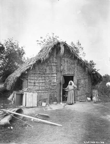 View across yard towards a woman standing at the open door of a grass hut. There are chickens around her in the yard, and inside the hut.