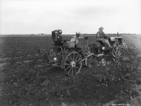 Three-quarter view from right rear of two men working in a field. One man is driving a Farmall tractor which is pulling another man who is sitting on a potato digger. There are buildings in the distance