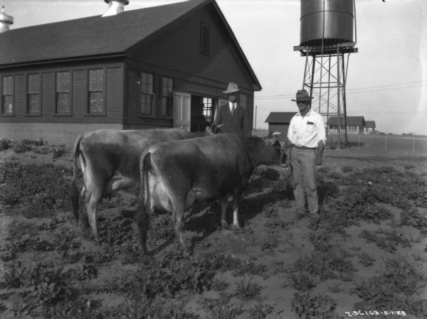 Two men are standing in a barnyard with two cows. Behind them on the left is a large barn, and in the background is a water tower and other buildings.