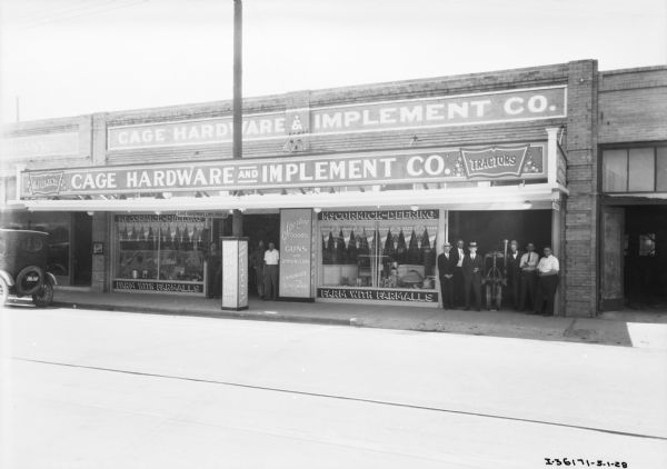 View across street towards a group of people standing at the entrance to the Cage Hardware and Implement Co. in the center, and another group of peoplestanding at the open garage entrance on the right with a tractor.