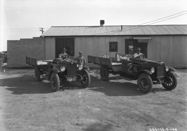 Two men are sitting in the driver's seats of two trucks with open truck beds. The trucks have a hood ovr the engine, but the driver's seat is exposed. Another man is standing near the truck on the left, and a building in the background has a sign that reads: "R.W. Briggs & Co. Contractors, Pharr, Texas."