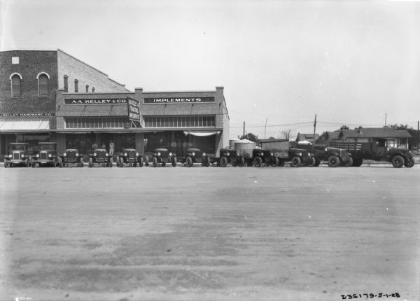 View across street towards the A.A. Kelley & Co. Implements and Hardware store. Trucks are lined up along the sidewalk, and groups of men are posing at two of the entrances.