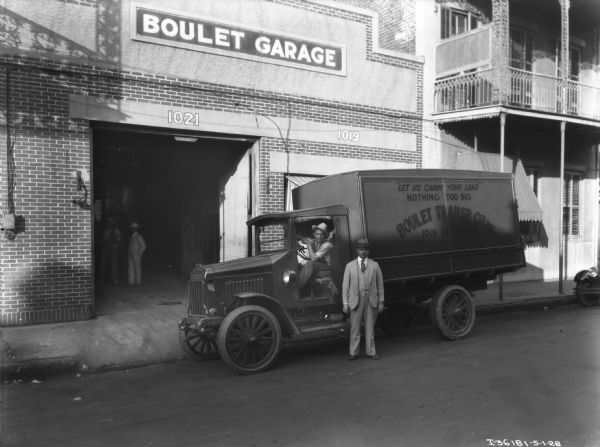 A man is sitting in the driver's seat of a truck parked in front of the garage entrance door of a brick building. Another man is standing in the street near the truck driver. More men are standing inside of the garage. The sign painted on the truck reads: "'Let Us Carry Your Load' Nothing Too Big, Boulet Transp. Co. Inc., 1019 St. Louis St."