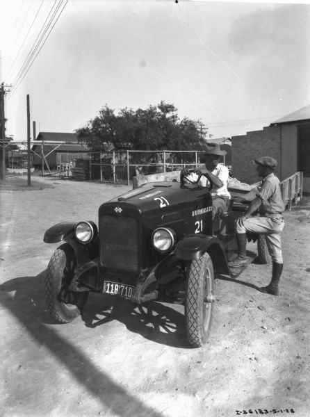 One man is sitting in the driver's seat of the truck, and the other man is standing at the side with one foot on the running board. Industrial buildings are in the background.