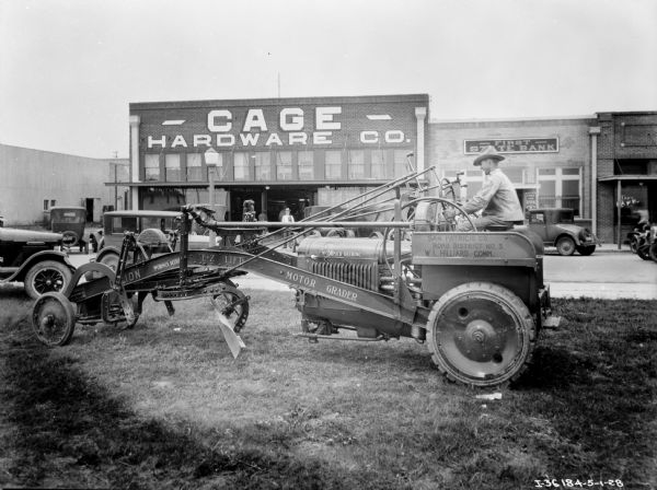 Left side profile view of a man operating a McCormick-Deering tractor with a front loader. In the background are parked automobiles, and the Cage Hardware Co., and First State Bank buildings.