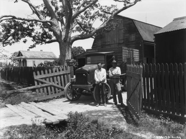 Two men are posing standing in front of a truck parked near a gate in a fence. There is a large tree, and a barn in the background.
