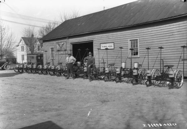 View towards two men standing near a row of planters in front of a dealership. A sign on the wall reads: "Farm Machine Headquarters, McCormick-Deering, Gaithersburg, Md."