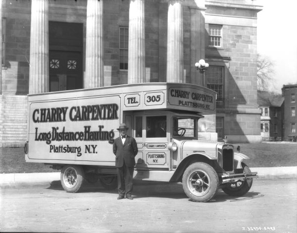 View towards a man standing in front of a C. Harry Carpenter truck. "Long Distance Hauling." A man is sitting in the driver's seat of the truck, which is parked in front of a large, stone building with tall columns.