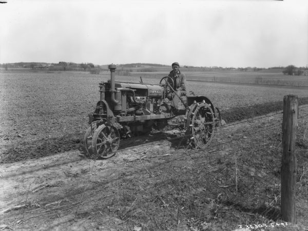 Three-quarter view from front left of a man using a Farmall tractor for plowing a field. There is a fence in the foreground.