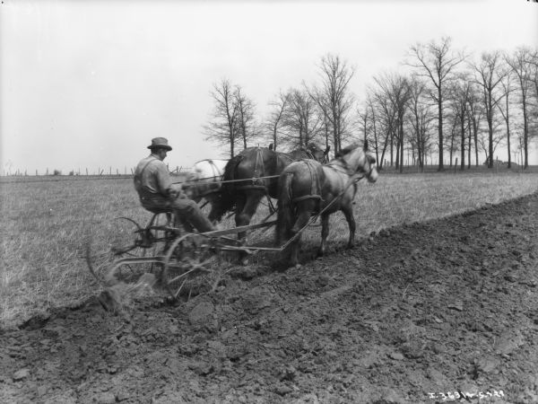 Three-quarter view from right rear of a man on a horse-drawn plow in a field.