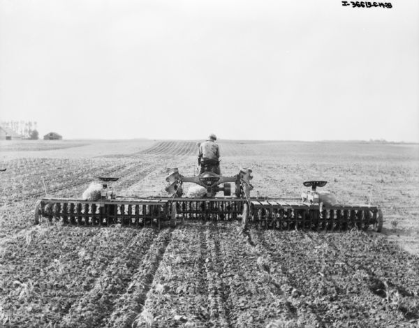 Rear view of a man using a Farmall tractor to pull three rotary hoes in a field.