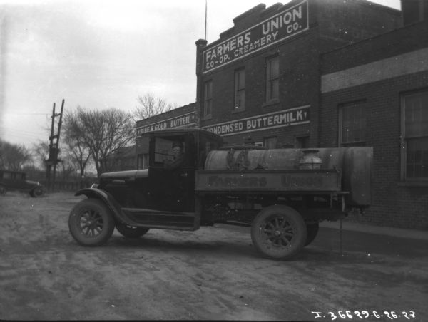 View of a man sitting in the driver's seat of a Farmers Union truck. In the background is the Farmers Union dairy building.
