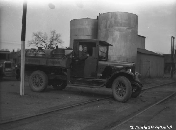 View towards a man sitting in the driver's seat of a Farmers Union delivery truck at the dairy. Storage tanks are in the background.